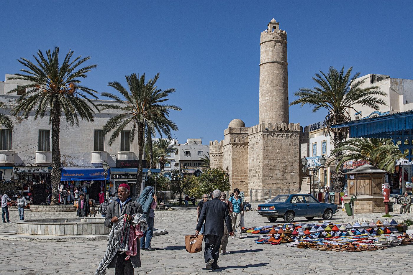 People walking past a market stall in the sunny medina square below the Ribat fortress and tower in Sousse